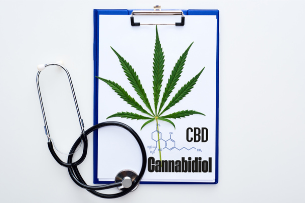 bovenaanzicht van medisch cannabisblad op klembord met cbd letters bij stethoscoop op witte achtergrond - Foto, afbeelding