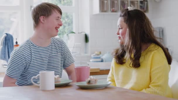 Young Downs Syndrome couple sitting around table with hot drink and cakes at home talking - shot in slow motion - Imágenes, Vídeo
