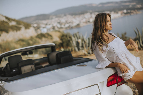 Mulher bonita pelo carro branco do cabriolet na beira-mar em um dia ensolarado do verão
 - Foto, Imagem