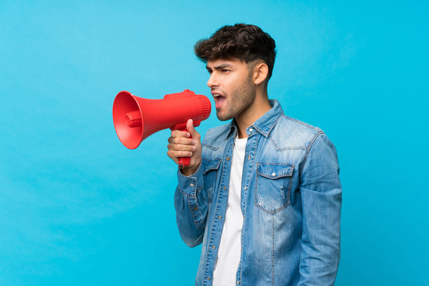 Молодой красивый мужчина на изолированном синем фоне кричит через мегафон
 - Фото, изображение