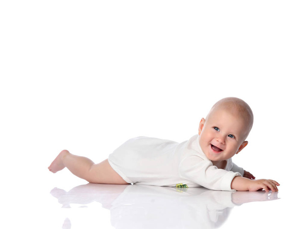 Smiling infant child baby toddler in white bodysuit lying on his stomach crawling, trying to reach something on white - Photo, Image