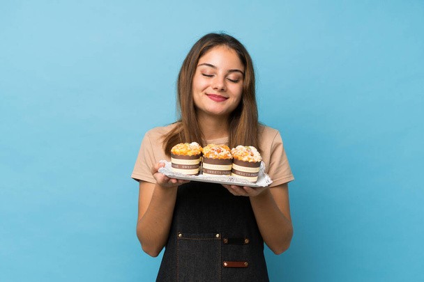 Genç esmer kız, izole edilmiş mavi arka planda elinde mini pastalarla onların kokusundan zevk alıyor. - Fotoğraf, Görsel