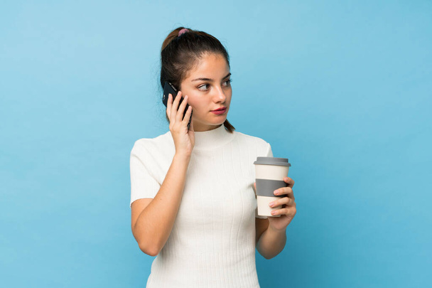 junge brünette Mädchen über isolierten blauen Hintergrund hält Kaffee zum Mitnehmen und ein Handy - Foto, Bild