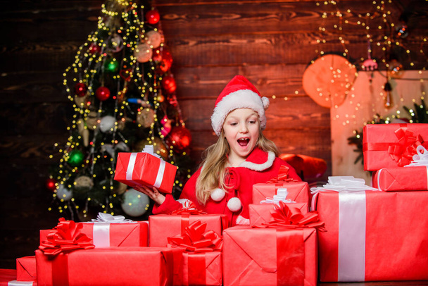 Child happy excited girl find gifts near christmas tree. Happiness and joy. Merry christmas. Happy childhood concept. Kid wear santa hat hold wrapped christmas gift box. Santa brought me gifts - Foto, immagini