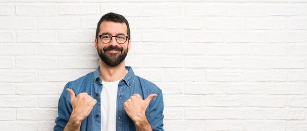 Handsome man with beard over white brick wall with thumbs up gesture and smiling - Valokuva, kuva
