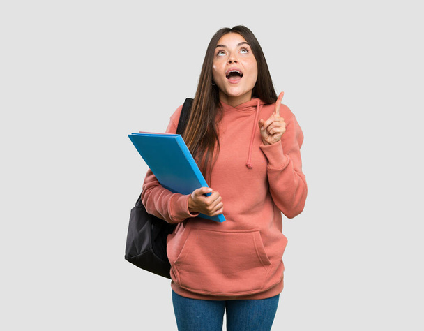 Joven estudiante sosteniendo cuadernos sorprendida y señalando hacia arriba sobre fondo gris aislado
 - Foto, imagen