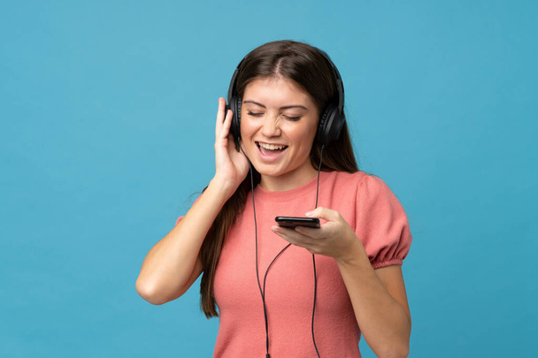 Young woman over isolated blue background using the mobile with headphones and singing - Photo, Image