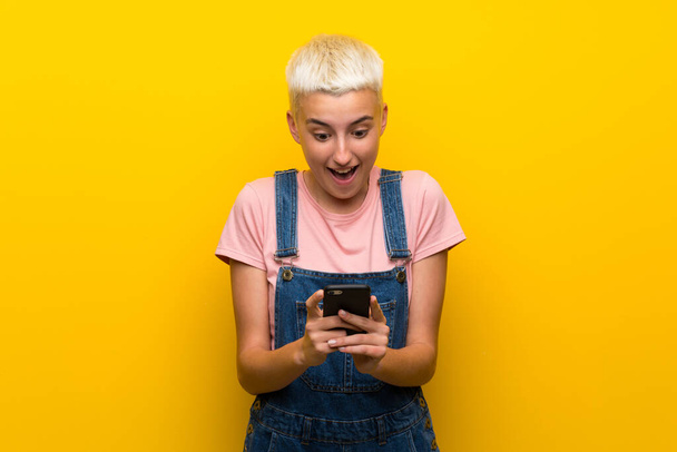 Teenager girl with overalls on yellow background surprised and sending a message - Foto, immagini