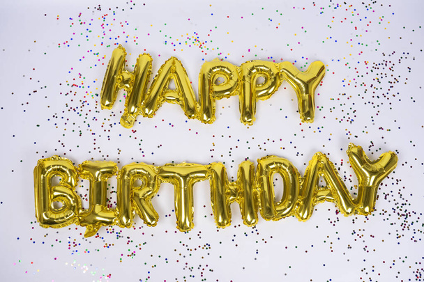 Gelukkige verjaardag belettering van gefrustreerde gouden ballonnen geïsoleerd met kleurrijke confetti op witte achtergrond. Gefeliciteerd.. - Foto, afbeelding