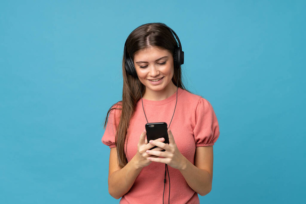 Jonge vrouw over geïsoleerde blauwe achtergrond met behulp van de mobiele telefoon met hoofdtelefoon - Foto, afbeelding