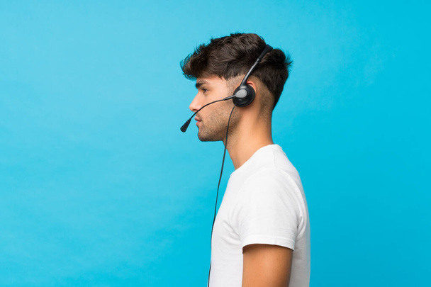 Νέος όμορφος άντρας πάνω από απομονωμένο μπλε φόντο που εργάζεται με ακουστικά - Φωτογραφία, εικόνα