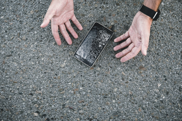 Σπασμένη οθόνη τηλεφώνου στο δρόμο. Ο άνθρωπος είναι δυσαρεστημένος. Οι άντρες έριξαν το τηλέφωνο από τα χέρια του στην άσφαλτο και έσπασαν την οθόνη. κινητό δεν είναι ανακτήσιμο - Φωτογραφία, εικόνα