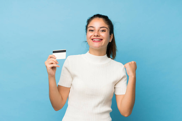 Young brunette girl over isolated blue background holding a credit card - Photo, Image