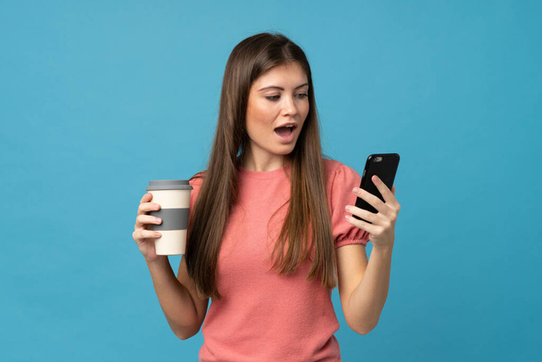 Young woman over isolated blue background holding coffee to take away and a mobile - Foto, Bild
