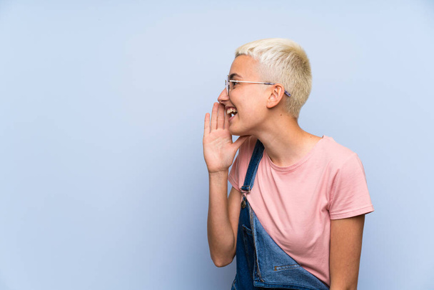 Teenager girl with overalls on blue wall shouting with mouth wide open - Photo, Image