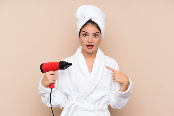 Young woman in a bathrobe with hairdryer over isolated background with surprise facial expression - Zdjęcie, obraz