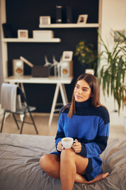 Charming brunette in blue sweater sitting in bedroom in morning and drinking coffee. - Photo, image