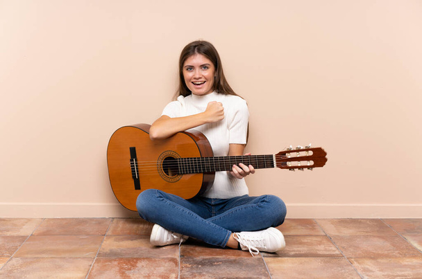 Νεαρή γυναίκα με κιθάρα κάθεται στο πάτωμα γιορτάζοντας μια νίκη - Φωτογραφία, εικόνα