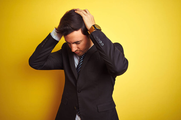Young handsome businessman wearing suit and tie standing over isolated yellow background suffering from headache desperate and stressed because pain and migraine. Hands on head. - Zdjęcie, obraz