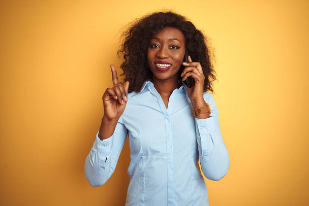 Genç Afro-Amerikalı kadın, sarı arka planda, akıllı telefondan konuşuyor. Bir fikir ya da soru işareti ile şaşırmış, mutlu yüzlü, bir numara. - Fotoğraf, Görsel