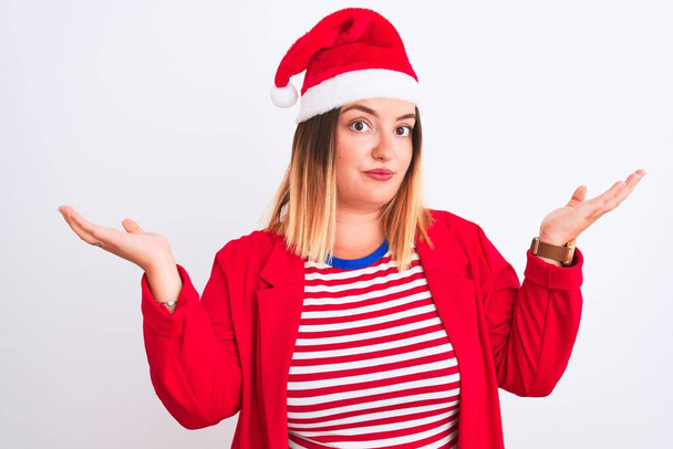 Young beautiful woman wearing Christmas Santa hat over isolated white background clueless and confused expression with arms and hands raised. Doubt concept. - Foto, imagen
