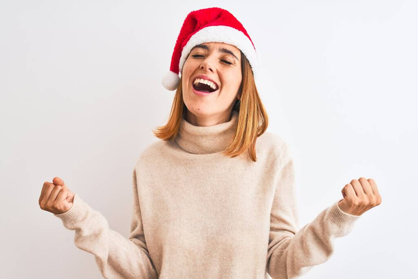Красивая рыжая женщина в рождественской шляпе на изолированном фоне празднует удивление и удивление за успех с поднятыми руками и закрытыми глазами. Концепция победителя
. - Фото, изображение