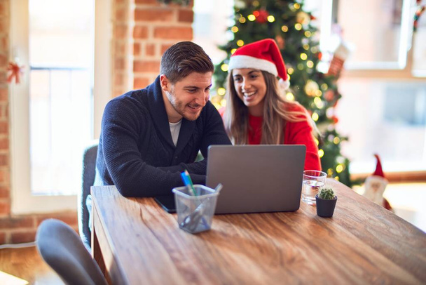 若い美しいカップルの幸せと自信を笑顔。自宅でクリスマスツリーの周りのノートパソコンを使用して机の上に座って - 写真・画像