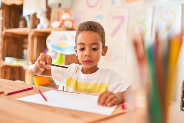 Piękny afrykański maluch siedzi rysunek przy użyciu papieru i ołówków na biurku w przedszkolu - Zdjęcie, obraz