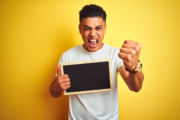 Young brazilian man holding blackboard standing over isolated yellow background annoyed and frustrated shouting with anger, crazy and yelling with raised hand, anger concept - Photo, Image