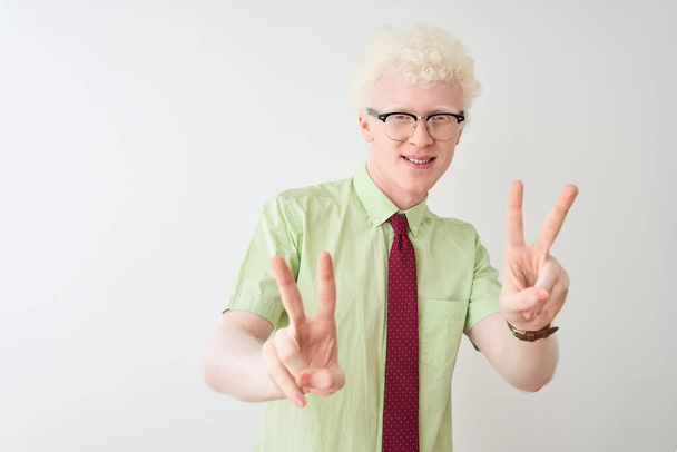 Der junge Albino-Geschäftsmann mit Hemd und Krawatte steht lächelnd vor isoliertem weißem Hintergrund und schaut in die Kamera, wie er mit den Fingern ein Siegeszeichen macht. Nummer zwei. - Foto, Bild