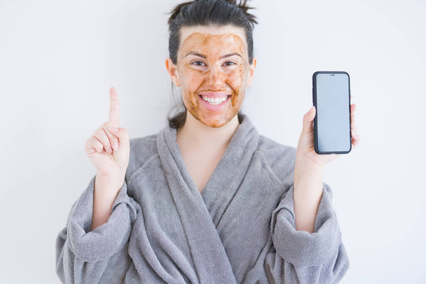 Красивая женщина носит косметическую маску для лица в качестве лечения по уходу за кожей, показывая экран смартфона, удивленный идеей или вопросом, указывая пальцем со счастливым лицом, номер один
 - Фото, изображение