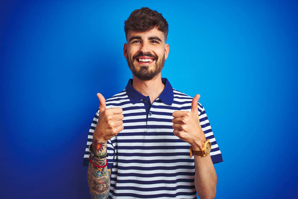 孤立した青の背景の成功のサインの上に立ってストライプポロを身に着けている入れ墨を持つ若い男は、手で積極的なジェスチャーを行います,親指を笑顔と幸せ.陽気な表情と勝者のジェスチャー. - 写真・画像