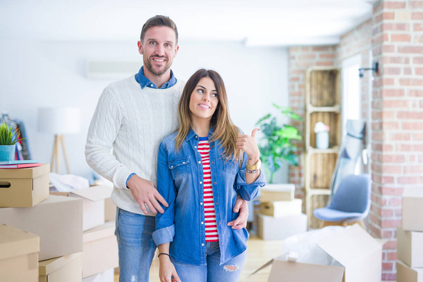 Young beautiful couple standing at new home around cardboard boxes smiling with happy face looking and pointing to the side with thumb up. - Photo, Image