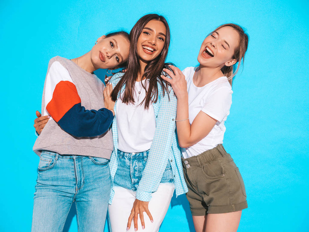 Three young beautiful smiling hipster girls in trendy summer clothes. Sexy carefree women posing near blue wall in studio. Positive models having fun and hugging - Fotografie, Obrázek