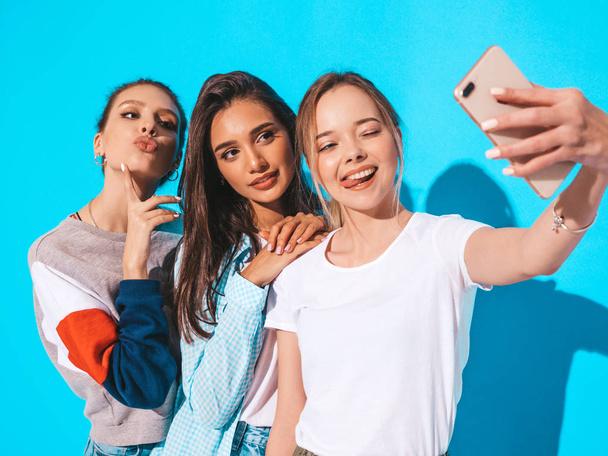 Три молодые улыбающиеся хипстерши в летней одежде. Девушки делают селфи с автопортретом на смартфоне. Модели позируют возле голубой стены в студии.
 - Фото, изображение