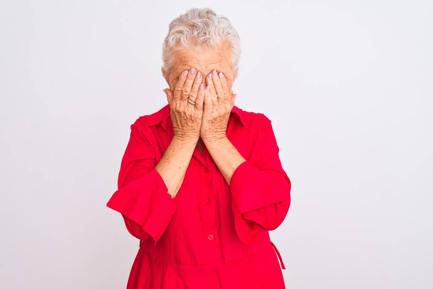 Idősebb, ősz hajú nő, piros alkalmi ingben, elszigetelt fehér háttér fölött áll, szomorú arckifejezéssel, miközben kezekkel takarja el az arcát, miközben sír. Depressziós koncepció. - Fotó, kép