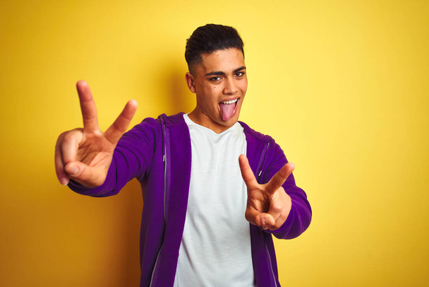 紫のスウェットシャツを着た若いブラジル人男性が孤立した黄色の背景の上に立っていて、両手の指が勝利サインをしているのを舌で笑っている。第二番. - 写真・画像