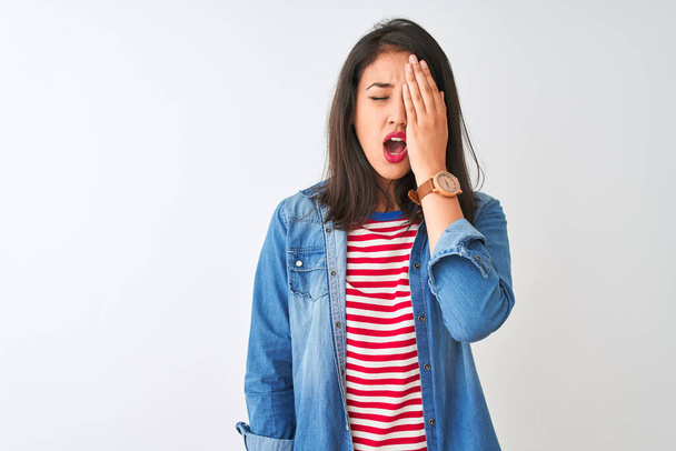 junge Chinesin in gestreiftem T-Shirt und Jeanshemd über isoliertem weißem Hintergrund gähnend müde, das halbe Gesicht, Augen und Mund mit der Hand verdeckt. Gesicht schmerzt vor Schmerzen. - Foto, Bild