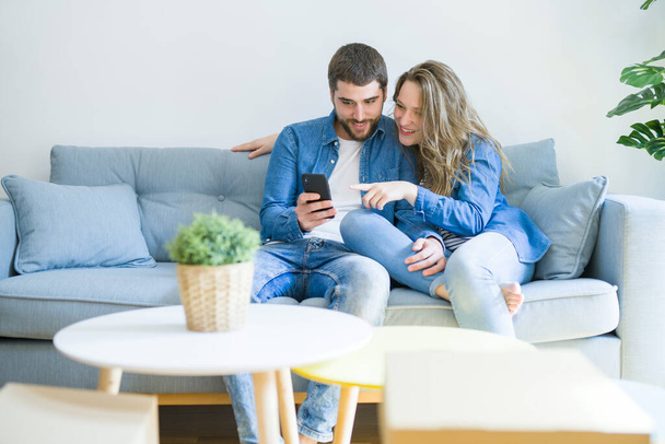 Jeune couple amoureux étreignant assis sur le canapé à l'aide d'un smartphone très heureux de déménager dans une nouvelle maison
 - Photo, image