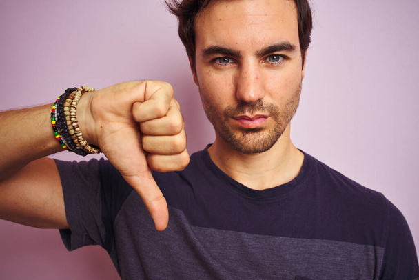 Young handsome man wearing casual t-shirt standing over isolated pink background with angry face, negative sign showing dislike with thumbs down, rejection concept - Photo, Image