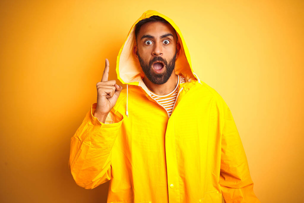 Nuori intialainen mies yllään sadetakki huppu seisoo yli eristetty keltainen tausta osoittaa sormella ylös onnistunut idea. Poistunut ja onnellinen. Numero yksi.
. - Valokuva, kuva