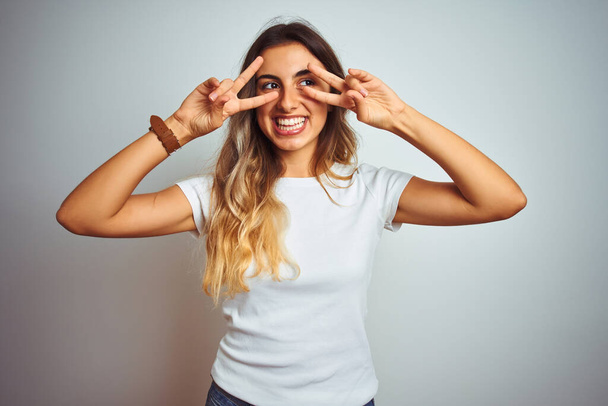 Young beautiful woman wearing casual white t-shirt over isolated background Doing peace symbol with fingers over face, smiling cheerful showing victory - Фото, зображення