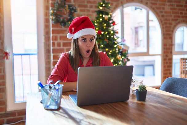 Hermosa mujer sentada a la mesa trabajando con portátil con sombrero de Santa Claus en Navidad asustada y sorprendida con la expresión sorpresa, el miedo y la cara emocionada
. - Foto, imagen
