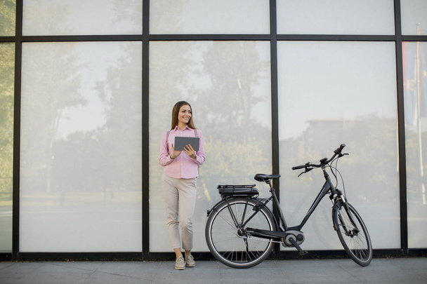 Mujer bastante joven de pie junto a una bicicleta eléctrica y el uso de tabletas digitales en el entorno urbano
 - Foto, Imagen
