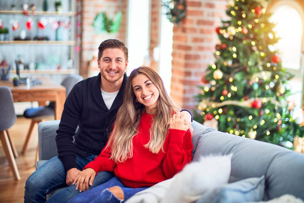 Young beautiful couple smiling happy and confident. Sitting on the sofa hugging around christmas tree at home - Fotoğraf, Görsel