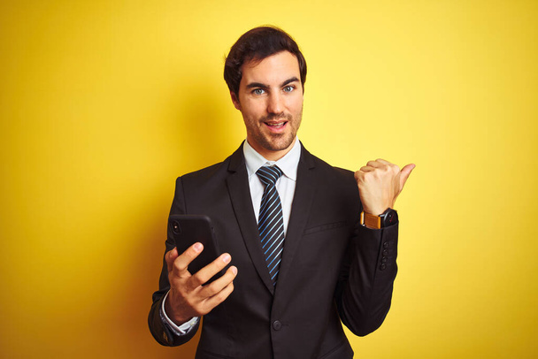 Молодий красивий бізнесмен використовує смартфон, що стоїть над ізольованим жовтим тлом, вказуючи і показуючи з великим пальцем в сторону з щасливим обличчям посміхаючись
 - Фото, зображення