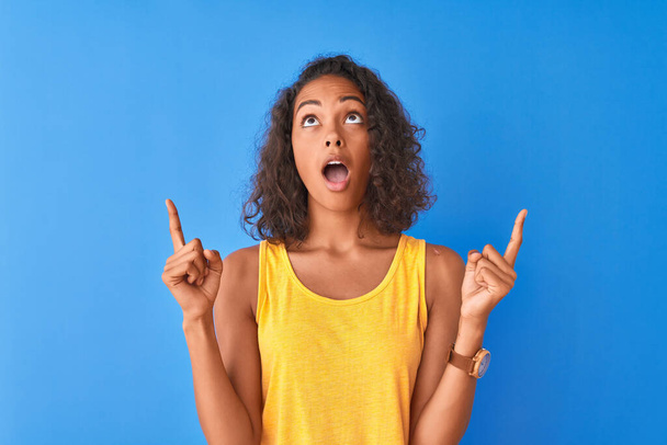 Young brazilian woman wearing yellow t-shirt standing over isolated blue background amazed and surprised looking up and pointing with fingers and raised arms. - Fotó, kép