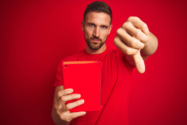 Fiatal férfi használ digitális touchpad tabletta felett piros elszigetelt háttér dühös arc, negatív jel azt mutatja, nem tetszik hüvelykujjal lefelé, elutasítás koncepció - Fotó, kép