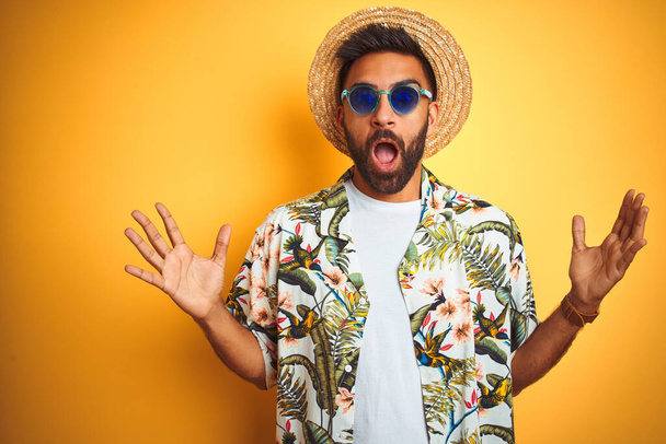 Индиец в отпуске в очках в шляпе из цветочной рубашки на изолированном желтом фоне празднует безумие и удивляется успеху с поднятыми руками и открытыми глазами, крича взволнованно. Концепция победителя
 - Фото, изображение