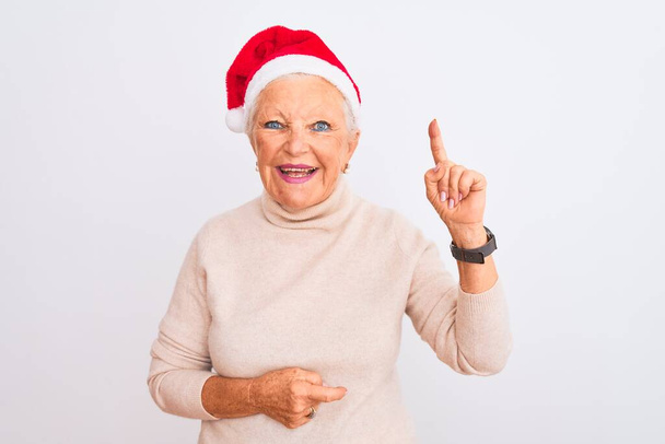 Senior harmaa tukka nainen yllään Crhistmas Santa hattu yli eristetty valkoinen tausta osoittaa sormella ylös onnistunut idea. Poistunut ja onnellinen. Numero yksi.
. - Valokuva, kuva